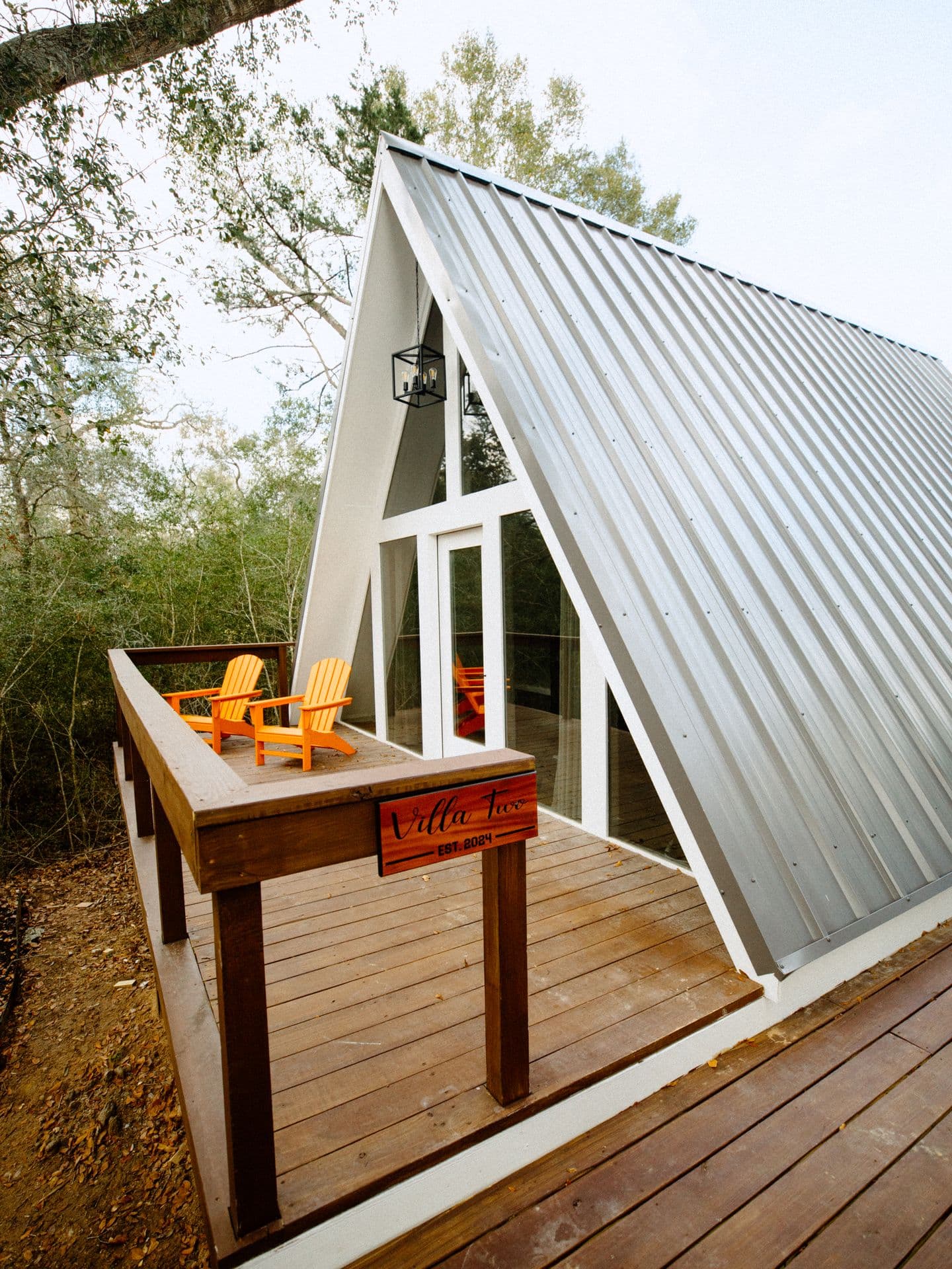 An A-frame cabin with a wraparound deck and orange chairs among the trees