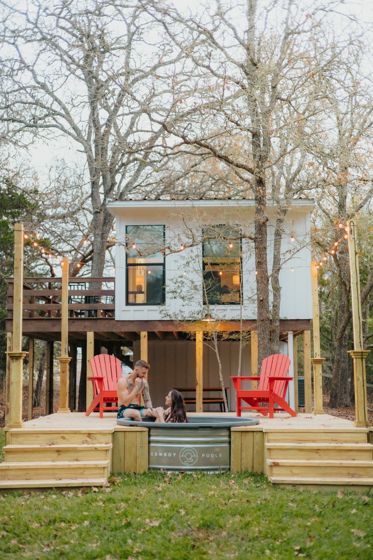 A couple in a stock-tank hot tub on the deck below the treehouse, string lights above